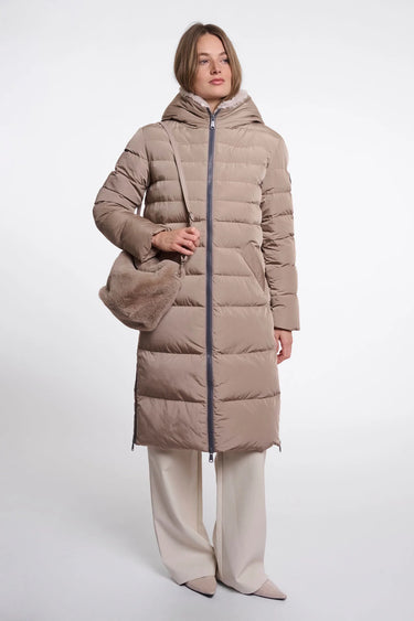 A woman modeling a long taupe-beige hooded puffer coat with a dark zipper, paired with light beige wide-leg trousers and a matching taupe shoulder bag, against a plain light gray studio background.