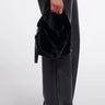 A person wearing black wide-leg leather pants, a black fuzzy top, and black pointed shoes holds a black fuzzy handbag with a small black pouch attached, photographed against a light gray studio background.