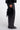 A person wearing black wide-leg leather pants, a black fuzzy top, and black pointed shoes holds a black fuzzy handbag with a small black pouch attached, photographed against a light gray studio background.