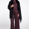 A woman modeling a black fringed faux-fur cape over a matching dark plum satin top and wide-leg trousers, photographed against a light gray studio background.