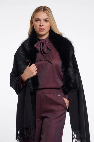 A blonde model wearing a black fringed faux-fur cape over a matching dark plum satin blouse and trousers, photographed against a plain light gray studio background.