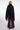 A woman shown from the back wearing a black shawl with dark fur trim over a burgundy wide-leg dress, photographed against a light gray studio background.