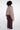 A model shown from the back wearing a dusty pink draped wrap or poncho over a brown faux-fur collar, paired with dark burgundy wide-leg trousers, against a plain white studio background.