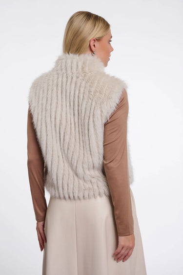 A woman shown from the back wearing a cream-colored faux fur vest over a brown long-sleeve top and a light beige skirt, against a plain white studio background.