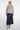 A model stands against a light gray studio background wearing a light beige cropped faux-fur jacket with a high collar and three front buttons, paired with a dark navy midi skirt and beige ankle boots.