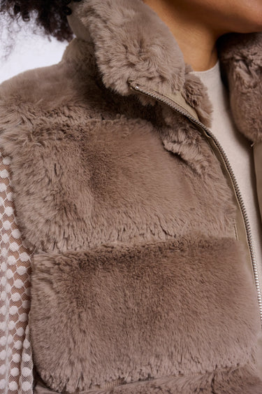 Close-up of a taupe faux-fur vest with a high collar and a silver zipper, worn over a white top.