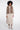 A model wearing a taupe faux fur vest with sheer patterned sleeves, a beige midi skirt, and beige ankle boots, photographed against a seamless white studio background.