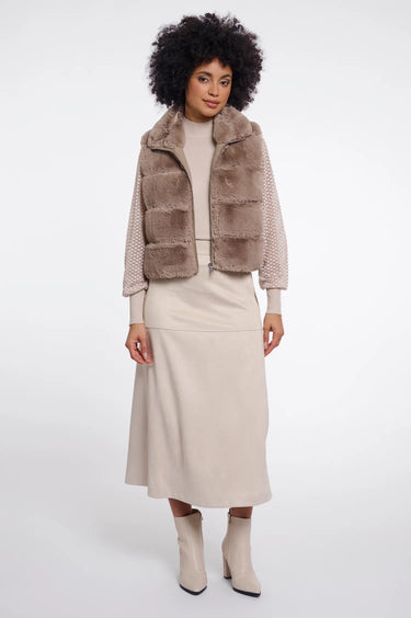 A model wearing a taupe faux fur vest with sheer patterned sleeves, a beige midi skirt, and beige ankle boots, photographed against a seamless white studio background.