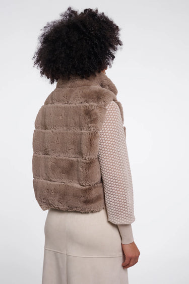A model shown from the back wearing a taupe-brown faux fur puffer vest over a beige knit sweater and cream skirt, against a plain white studio background.