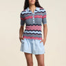 A woman wearing a zig zag stripe polo in multi color with blue shorts on a neutral background