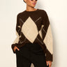 A woman wearing a brown and cream argyle knit sweater with black trousers, styled with gold-framed sunglasses, standing against a plain off-white wall.