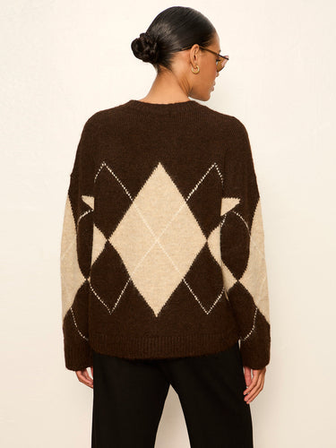 A woman viewed from the back wearing a brown and cream argyle knit sweater and black pants, standing against a plain off-white wall.