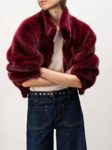 A model wearing a deep burgundy faux fur cropped jacket over a cream top and dark blue jeans, shown from the neck down against a plain light background.