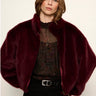 A woman wearing a plush, cropped burgundy faux fur jacket over a sheer black top and black pants, photographed against a plain off-white background.