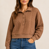 A woman wearing a brown cable-knit polo sweater with a button placket and blue jeans, standing against a plain beige studio background.
