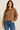 A woman wearing a brown cable-knit polo sweater with a button placket and blue jeans, standing against a plain beige studio background.