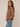 A woman wearing a brown cable-knit cropped sweater with a buttoned collar, paired with light blue jeans, standing against a plain beige studio background.
