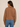 A woman shown from the back wearing a brown cable-knit cropped jacket with a collar and blue jeans, set against a plain beige studio background.