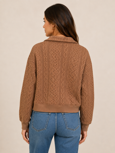 A woman shown from the back wearing a brown cable-knit cropped jacket with a collar and blue jeans, set against a plain beige studio background.