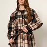 A woman modeling a long-sleeve belted plaid coat in brown, beige, and black tones, paired with dark brown leggings, against a plain light gray studio background.