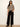 A woman modeling a beige and black horizontally striped short-sleeve knit top with black wide-leg pants and black ankle boots, standing against a plain off-white wall.