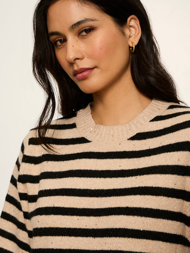 A close-up of a woman wearing a beige knit sweater with horizontal black stripes and small scattered sequins, paired with a gold hoop earring, against a plain light background.
