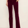 A person wearing deep burgundy corduroy pants with a slight flare at the hem, paired with black ankle boots, standing against a plain off-white wall.