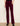 A person wearing deep burgundy corduroy pants with a slight flare at the hem, paired with black ankle boots, standing against a plain off-white wall.