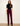 A woman stands against a plain off-white wall wearing a black short-sleeve mock-neck top and deep burgundy corduroy pants with a black studded belt, paired with black ankle boots.