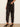 A model wearing black high-waisted satin trousers with side pockets and a cropped ankle length, styled with black slingback heels against a neutral cream wall and light wood floor.