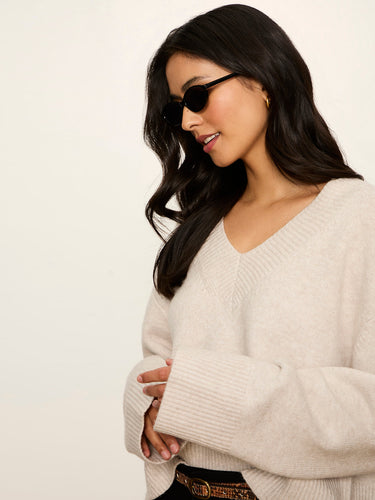 A woman with long dark hair wearing a cream-colored ribbed knit sweater, black sunglasses, and a gold hoop earring, shown in a side-profile view against a plain off-white background.