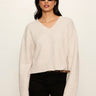 A woman modeling a cream-colored knit V-neck sweater with black pants and sunglasses, standing against a plain off-white background.