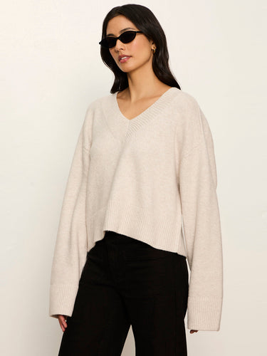 A woman modeling a light beige knit V-neck sweater with long sleeves, paired with black pants and black sunglasses, against a plain off-white background.