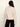 A woman viewed from the back wearing a cream-colored knit sweater and black pants, standing against a plain off-white background.