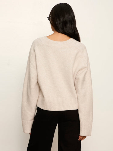 A woman viewed from the back wearing a cream-colored knit sweater and black pants, standing against a plain off-white background.
