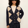 A model with curly black hair and sunglasses wearing a navy blue cardigan with a beige argyle pattern, styled with a cream top and dark jeans against a plain light background.