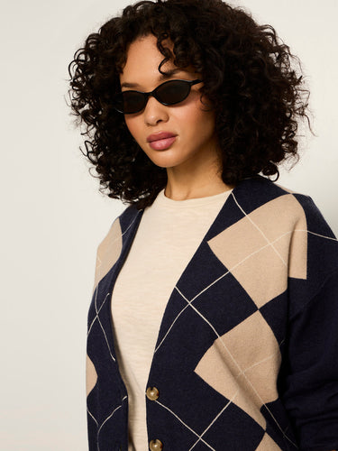 A woman with curly black hair wearing a navy and beige argyle cardigan over a cream top, styled with black sunglasses against a plain light background.