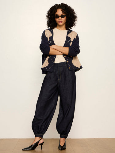 A model stands against a plain off-white wall wearing a dark navy and beige argyle cardigan, dark navy wide-leg denim pants, a cream top, and black pointed-toe heels.