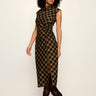 A woman modeling a sleeveless high-neck midi dress with a black and tan plaid pattern, paired with dark brown heeled boots, against a plain off-white studio background.