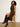 A woman with curly black hair sits on a wooden stool wearing a sleeveless black and tan plaid midi dress with a thigh-high slit and dark brown knee-high leather boots against a plain off-white wall.
