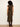 A woman viewed from behind wearing a sleeveless black midi dress with a gold plaid pattern and dark brown knee-high boots, standing against a plain off-white wall.