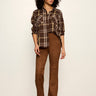 A model stands against a plain off-white wall wearing a brown and beige plaid button-up shirt with gold buttons and brown suede-like straight-leg pants.