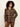 A woman with curly black hair wearing a brown and beige plaid button-up shirt and brown pants, standing against a plain off-white background.
