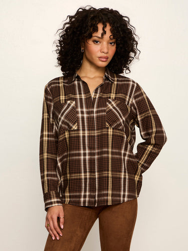A woman with curly black hair wearing a brown and beige plaid button-up shirt and brown pants, standing against a plain off-white background.