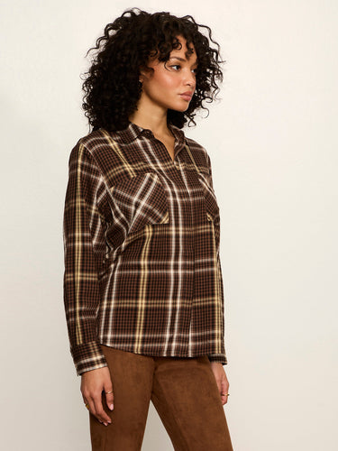 A woman with curly dark hair wearing a brown and beige plaid button-up shirt and brown pants, standing against a plain off-white background.