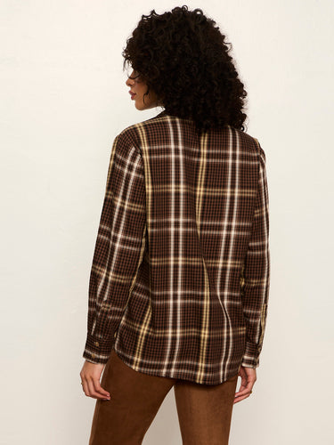 A woman with curly dark hair is shown from the back wearing a brown plaid button-up shirt with beige and white lines, paired with brown pants, against a plain off-white background.