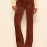 A person wearing brown corduroy flared pants with a black belt, paired with a cream lace long-sleeve top and black pointed-toe heels, standing against a plain light beige wall.