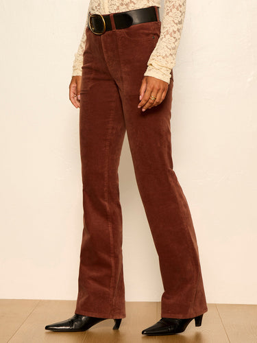 A person wearing brown corduroy flare pants with a black belt, paired with a cream lace long-sleeve top and black pointed-toe heels, standing against a plain off-white wall.