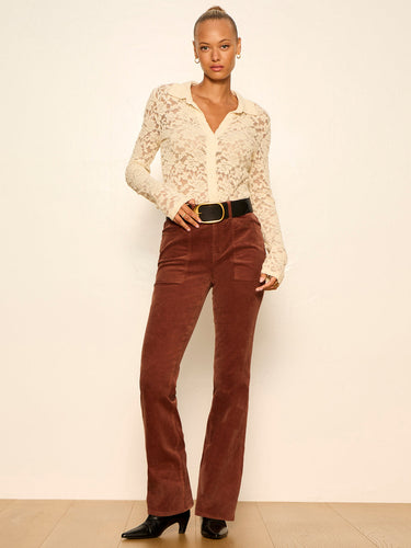 A model stands against a plain cream studio wall wearing a cream lace long-sleeve button-up shirt, brown corduroy flare pants with a black belt, and black pointed-toe heels.