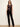 A woman modeling a sleeveless sequined top in a dark plum-brown color paired with black pants, standing against a plain off-white studio background.